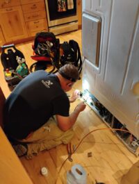 Technician servicing a refrigerator from below with tools laid out on the floor