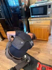 Technician kneeling behind a refrigerator to check the condenser fan