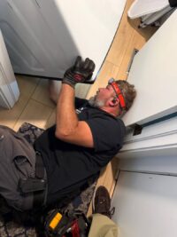 Technician on the floor with a headlamp checking under a washer for leaks